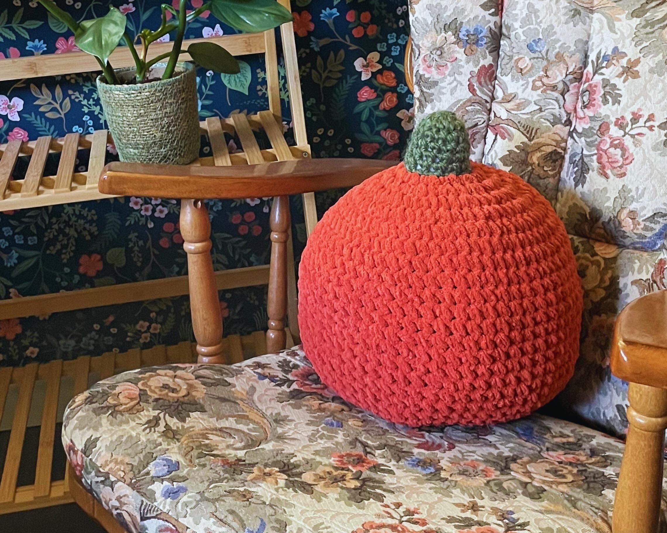 Orange crocheted pumpkin on a floral-patterned chair with a wooden stool and plant in the background.