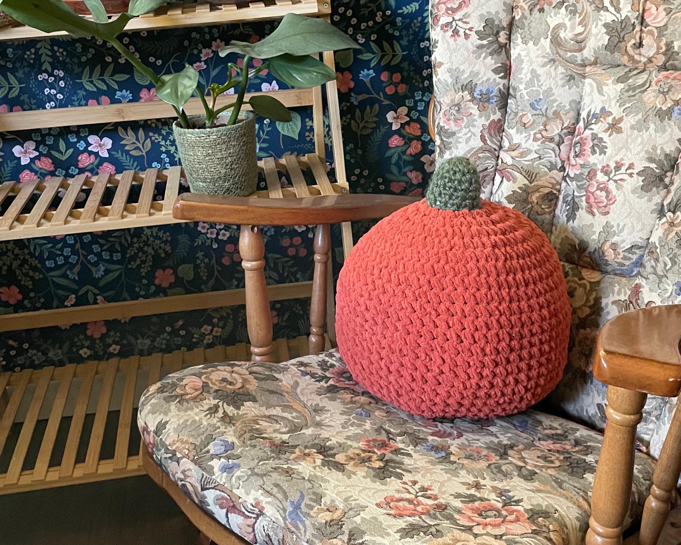 Crocheted orange pumpkin on a floral-patterned chair with a wooden shelf and plant in the background.